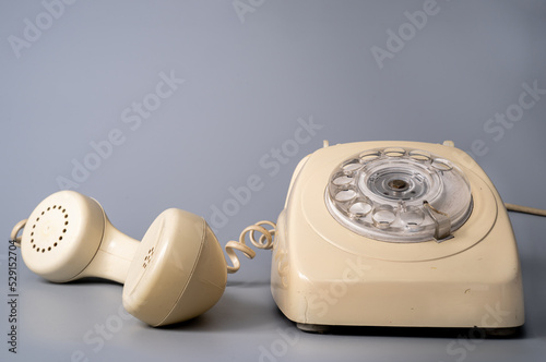 Old white rotary telephone with twisted cord on gray background. Retro landline phone with rotary dialer and remote handset. Vintage plastic rotary telephone with dialing disk.