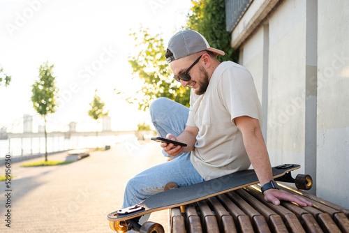 Happy young man with skateboard looking at mobile phone social media app to connect with friends. Urban lifestyle, street fashion and technology concept