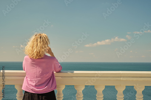 beautiful blonde adult woman in a pink t-shirt stands and enjoys a sea view from the balcony. Rear view, space for text