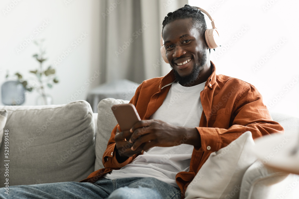 Excited african american man using smartphone while resting on couch at home, wearing wireless headset and smiling Excited african american man using smartphone while resting on couch at home, wearing wireless headset and smiling