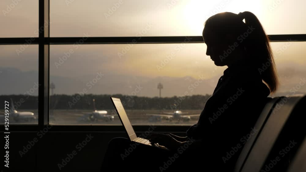 Freelancer lady does not waste time waiting for flight, uses every free minute to work, to complete urgent tasks. Silhouette of passenger sitting with laptop against lounge window. Blurred background