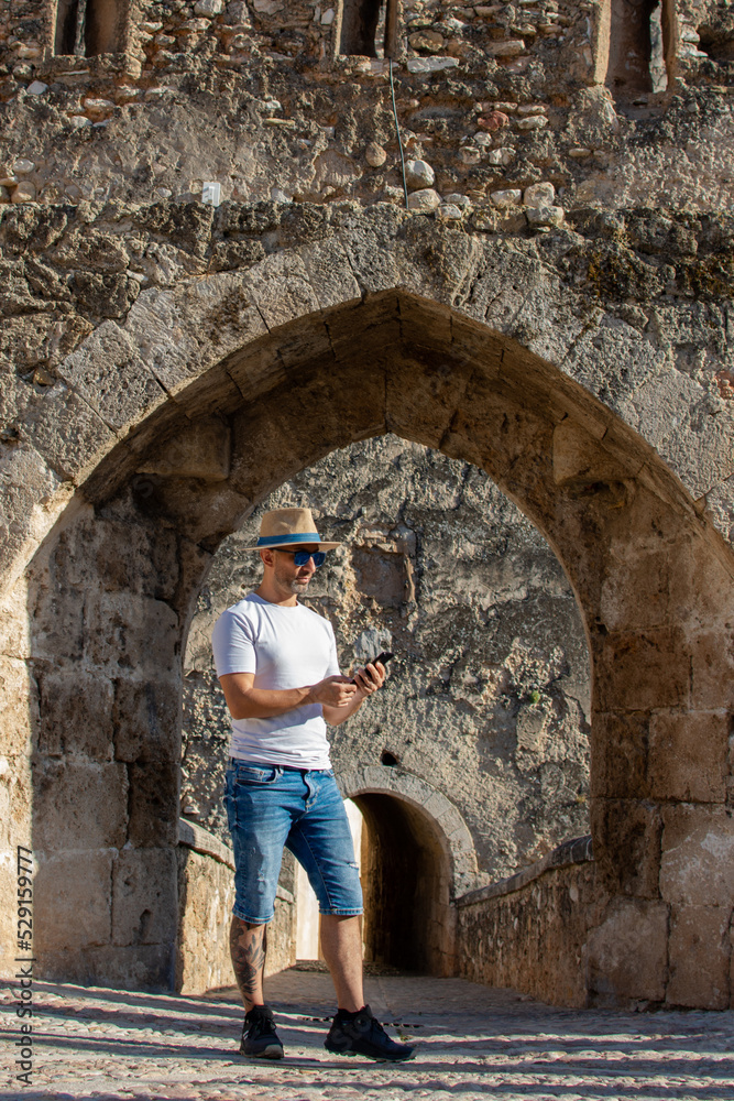 Man wearing hat and sunglasses standing in the doorway of an old stone castle while checking his cell phone