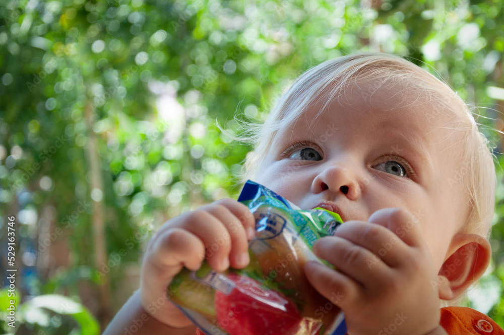 little blonde girl eats fruit puree in the park in summer, healthy baby food little blonde girl eats fruit puree in the park in summer, healthy baby food