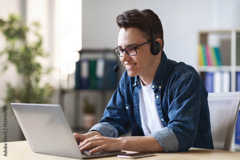 Fototapeta premium Telecommuting Concept. Young Male Manager In Headset Working With Laptop In Office