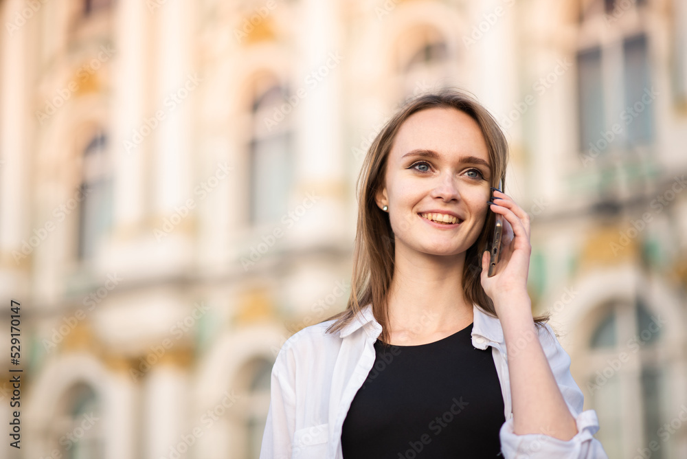 A young and attractive Caucasian girl in casual clothes smiles and talks on a mobile phone on a sunny day in the city. A young and attractive Caucasian girl in casual clothes smiles and talks on a mobile phone on a sunny day in the city.