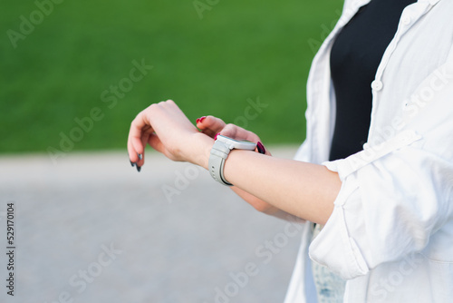 Close-up of a woman's hand in casual clothes using a smartwatch on her wrist in a park in nature. Checks the time on the clock. © Павел Костенко
