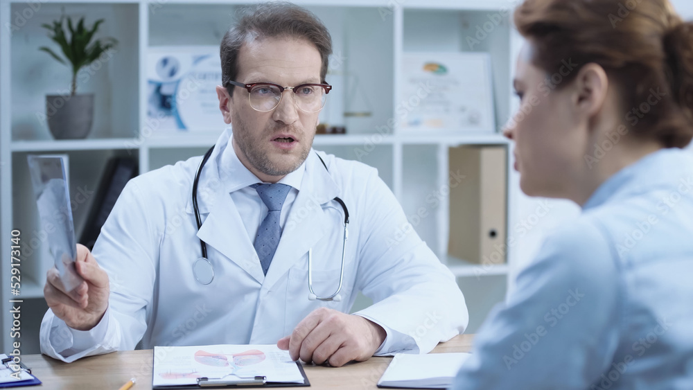 serious doctor holding x-ray scan while talking with patient on blurred foreground. serious doctor holding x-ray scan while talking with patient on blurred foreground.
