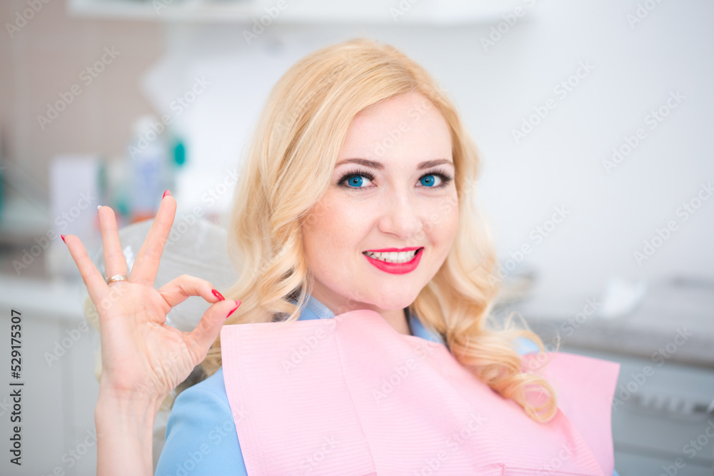 Dental clinic. A beautiful girl with a snow-white smile is sitting in the dental chair at the reception at the dentist. Dentistry and orthodontist. Dental treatment and bite correction. Sign is fine.