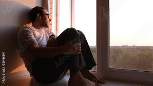 Depressed caucasian man losing job and heartbroken at same time alone, feeling bad and expresses negative. Unhappy attractive young male looks outside window in living room interior, close up.