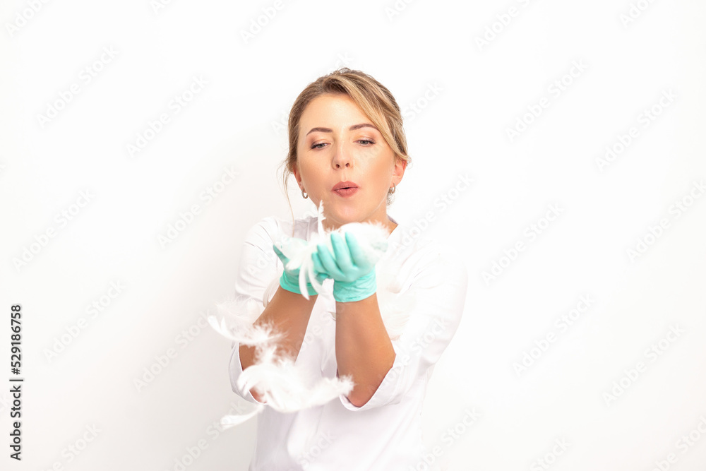 Young beautiful woman beautician in protective green gloves standing and blowing on feathers over white wall background