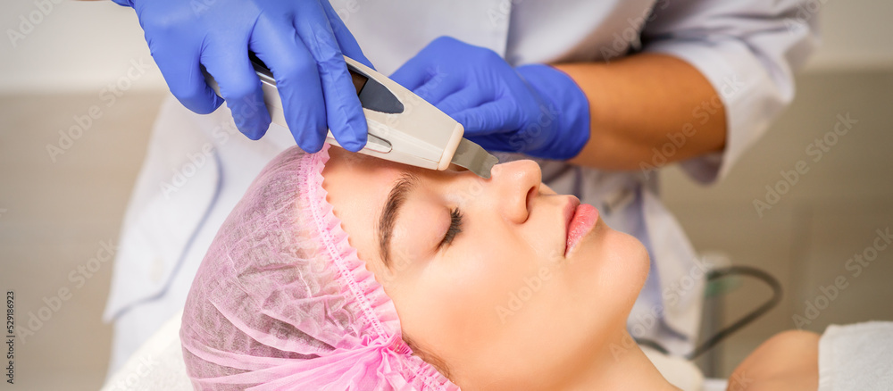 Naklejka premium Young caucasian woman receiving facial skin cleaning by ultrasonic cosmetology face equipment in a medical salon
