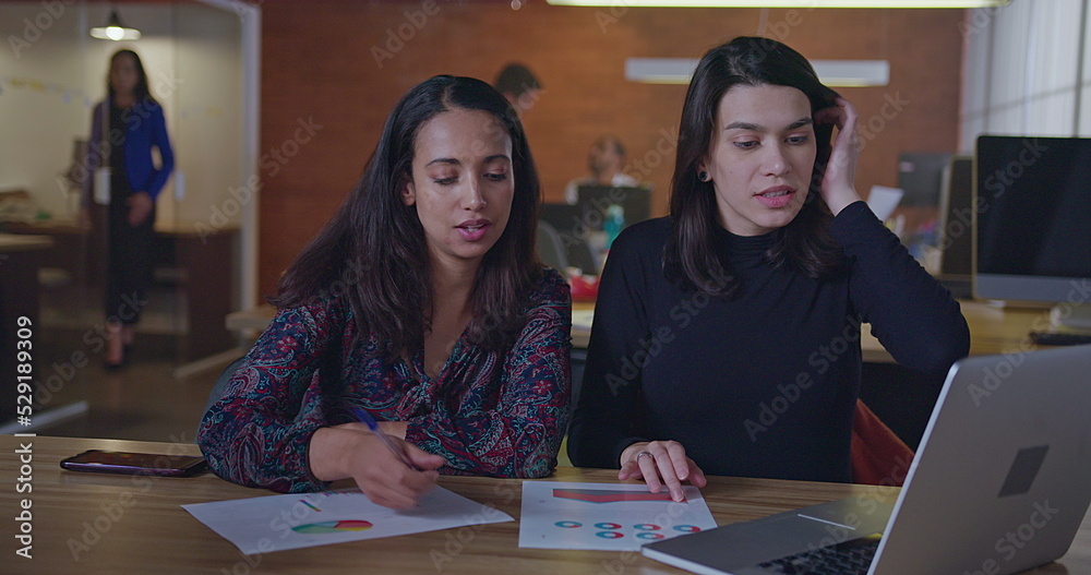 Two diverse female employees working at night in front of computer laptop sitting at office desk talking about work. Young women at their job inside modern workplace in the evening Two diverse female employees working at night in front of computer laptop sitting at office desk talking about work. Young women at their job inside modern workplace in the evening