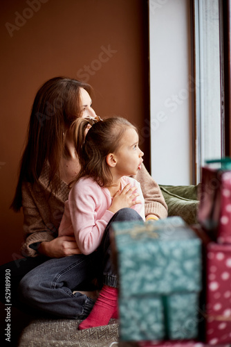 Little caucasian girl with mother looking at window and dreaming. A lot of gift boxes around. Happy family christmas concept
