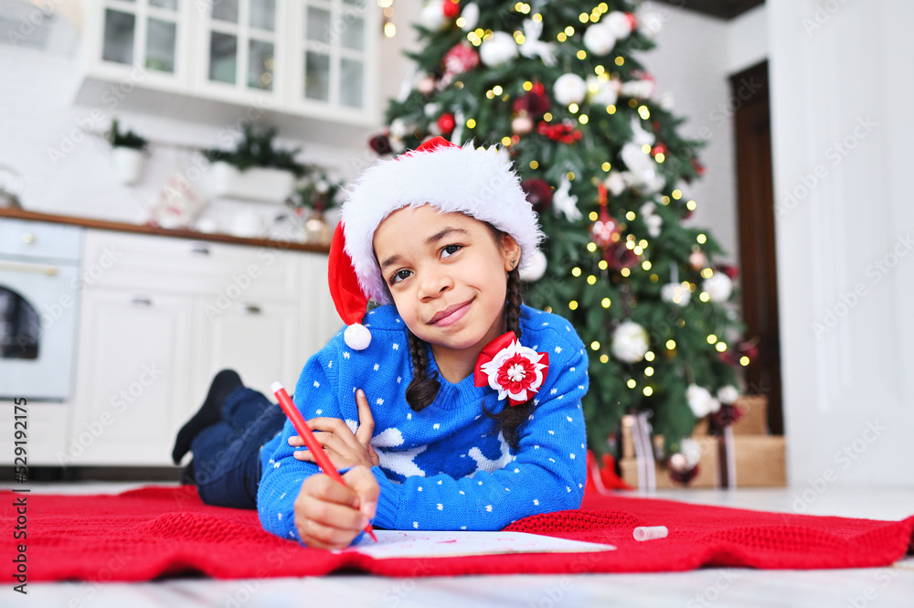 a little African-American girl in a blue sweater and a hat writes a wish letter to Santa Claus on the background of a Christmas tree.