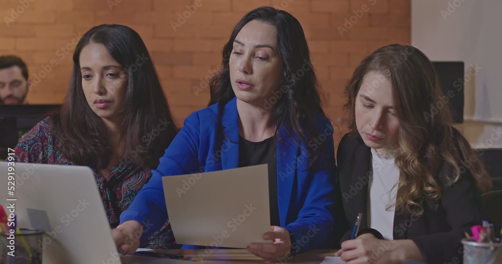 Women discussing work in front of laptop computer at corporate workplace. Female leader coaching younger female staff. People collaborating at night in the office Women discussing work in front of laptop computer at corporate workplace. Female leader coaching younger female staff. People collaborating at night in the office