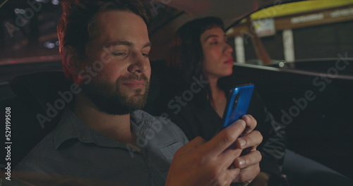 Young man showing cellphone screen to female work colleague in car backseat. People riding taxi looking at smartphone device. Person showing content online to friend