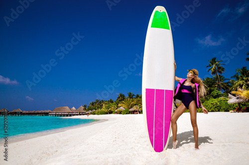 Beautiful sexy tanned woman in bikini is holding surfboard at Maldives sand beach. Maldives landscape. Girl with surfboard. Surfer girl. Luxury travel. Lifestyle. Tourism. Sport and recreation.