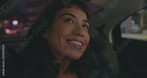 Happy female passenger in backseat of car smiling while looking out taxi window. Young woman visiting new city staring at buildings sightseeing urban street at night © Marco