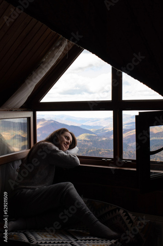 Young woman sitting in cozy house and looking at window. Mountain background
