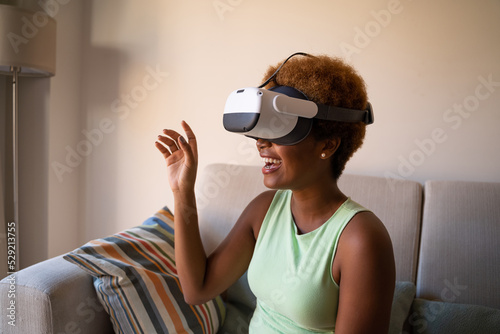 Smiling exited Black skin young woman wearing virtual reality Headset simulator playing VR game while sitting on sofa at home.  