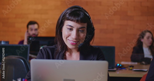 Confident young woman speaking with headset in front of computer screen at office call center. Female employee working at customer help desk talking