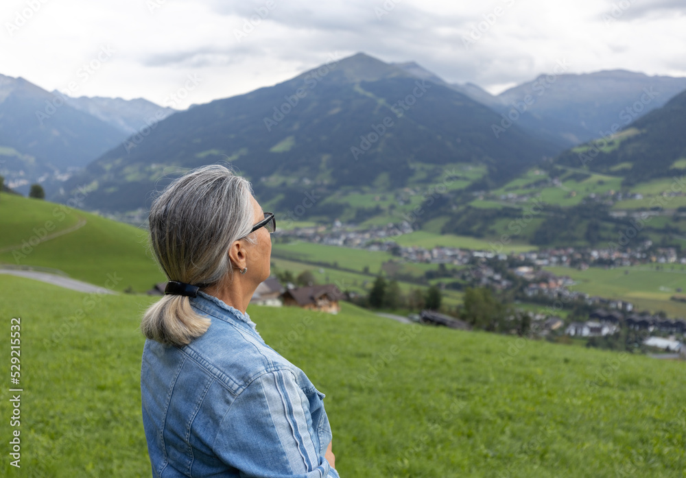A gray-haired woman in a denim suit stands near the road on a grassy slope and looks towards the mountains.