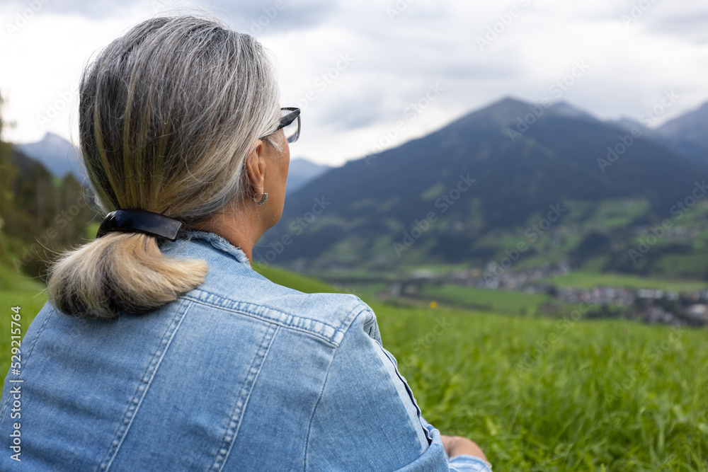 An elderly woman in a denim suit sits on a high meadow and looks at the mountains on the opposite side of the gorge.