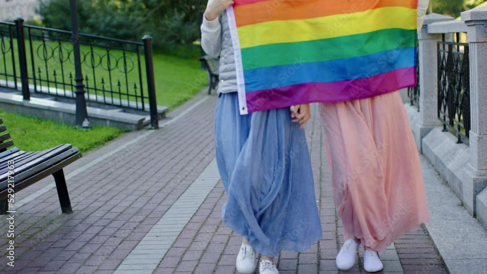 unrecognizable female couple walking outdoors with LGBT flag on camera ...
