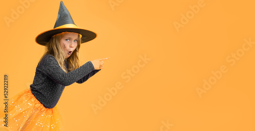 Excited kid girl wearing witch black hat costume Halloween isolated yellow studio background with finger pointing. Seasonal sale and discount. Shopping attentive. Banner, copy space for text, mock up