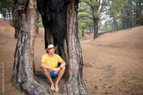 Hiker with hat  yellow shirt and blue shorts sits in the forest on ElHierro in a hollow burnt out tree and takes a break