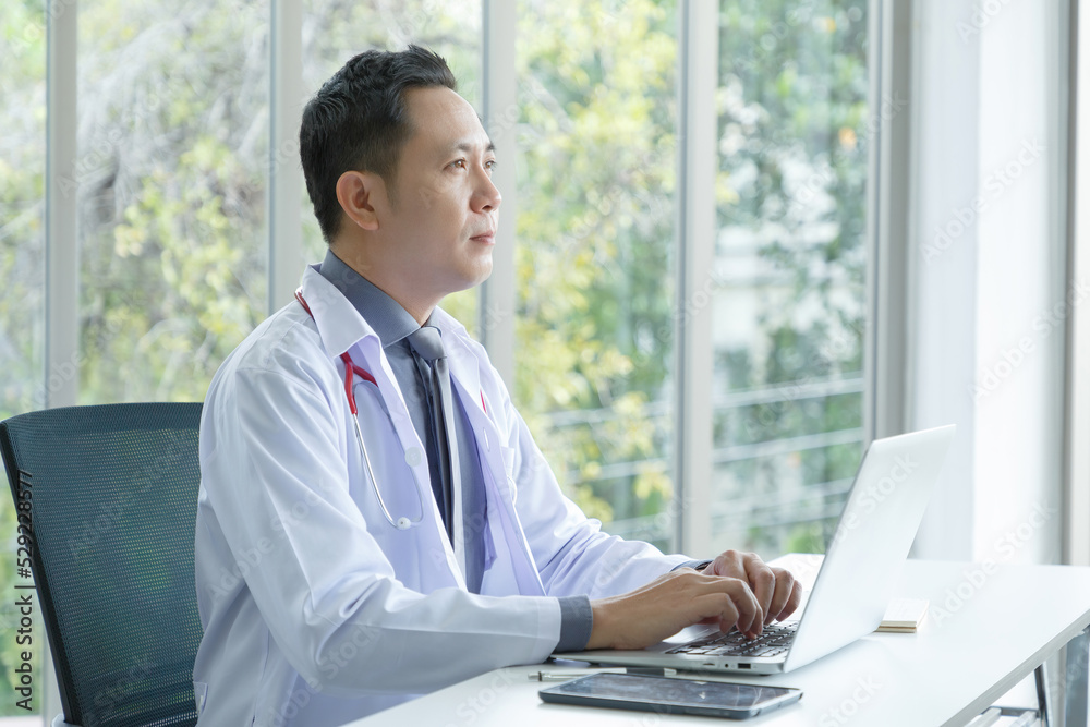 Portrait of asian senior doctor sitting in medical office while using laptop computer and checking time of appointment. Portrait of asian senior doctor sitting in medical office while using laptop computer and checking time of appointment.