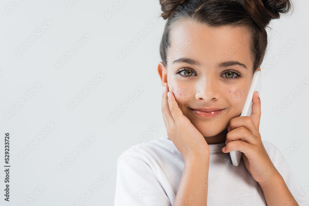 cheerful girl with glitter stars on cheeks talking on smartphone isolated on grey. cheerful girl with glitter stars on cheeks talking on smartphone isolated on grey.