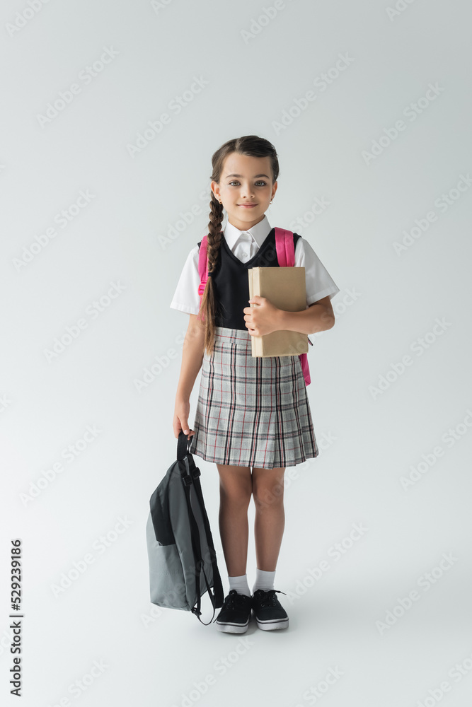 full length of adorable schoolgirl in uniform holding books and backpack on grey. full length of adorable schoolgirl in uniform holding books and backpack on grey.