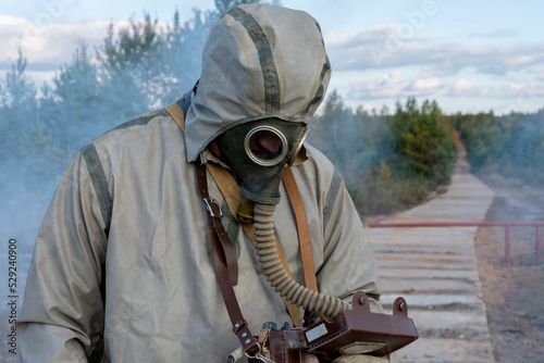 A military man in a chemical protection suit and a gas mask measures the level of radiation on a concrete road, smoke from fires. Concept: consequences of radiation leakage, photo