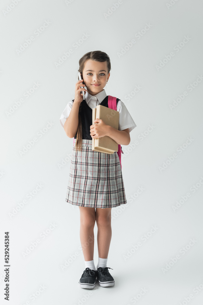 full length of smiling schoolgirl talking on smartphone and holding books on grey. full length of smiling schoolgirl talking on smartphone and holding books on grey.