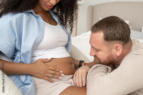 Handsome man father listens and look at pregnant tummy of his beautiful black wife and smiles. Multiracial marriage. Baby anticipation. Parenthood. Closeup