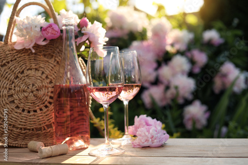 Fototapeta Naklejka Na Ścianę i Meble -  Bottle and glasses of rose wine near beautiful peonies on wooden table in garden
