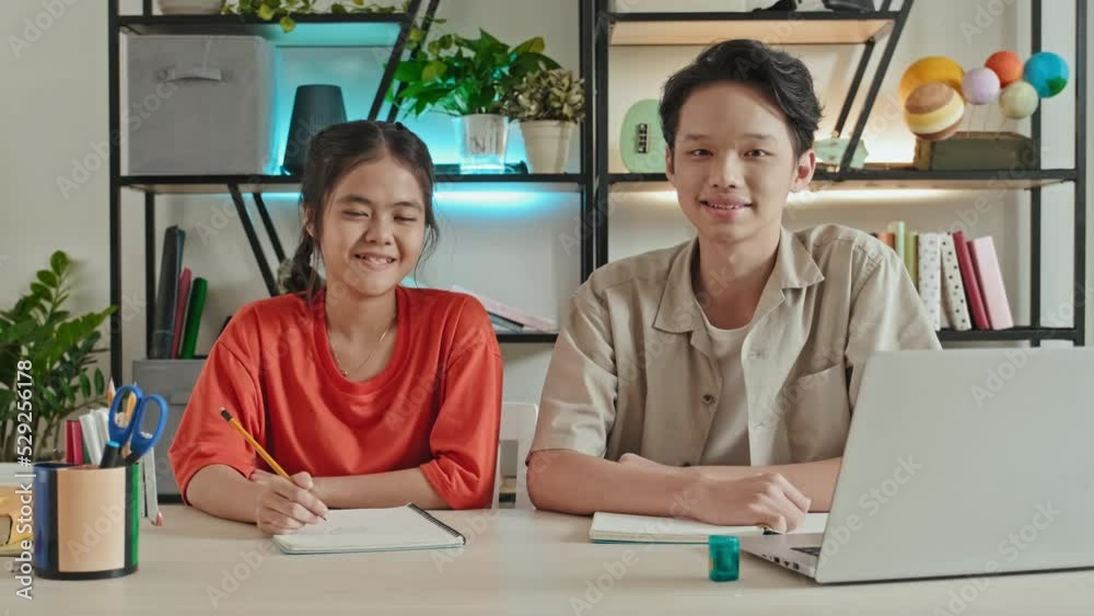 Portrait of happy Asian boy and girl sitting at desk in school classroom, looking at each other and then posing for camera with smile