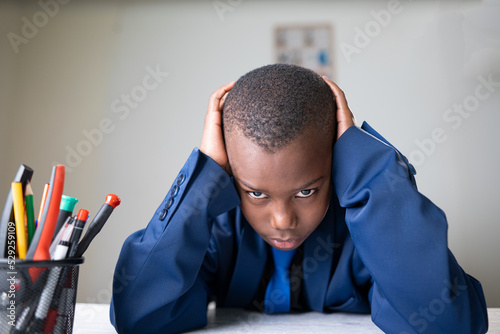 Giovane ragazzo nero in giacca e cravatta depresso seduta con i gomiti appogiate sulla scrivania e le mani intorno alla testa e guarda la telecamera  photo