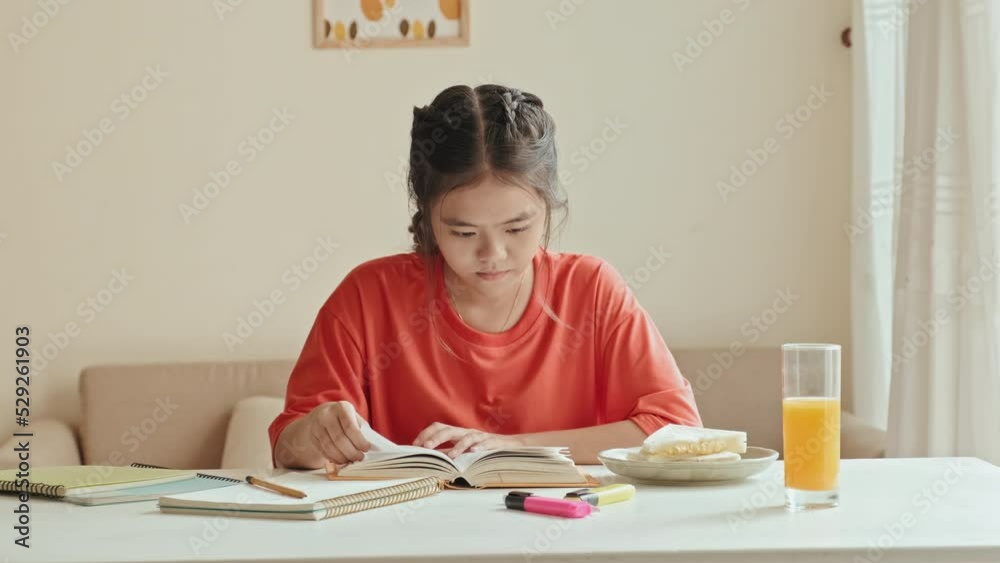 Asian girl sitting at table in living room, eating sandwich and reading textbook while doing homework Asian girl sitting at table in living room, eating sandwich and reading textbook while doing homework