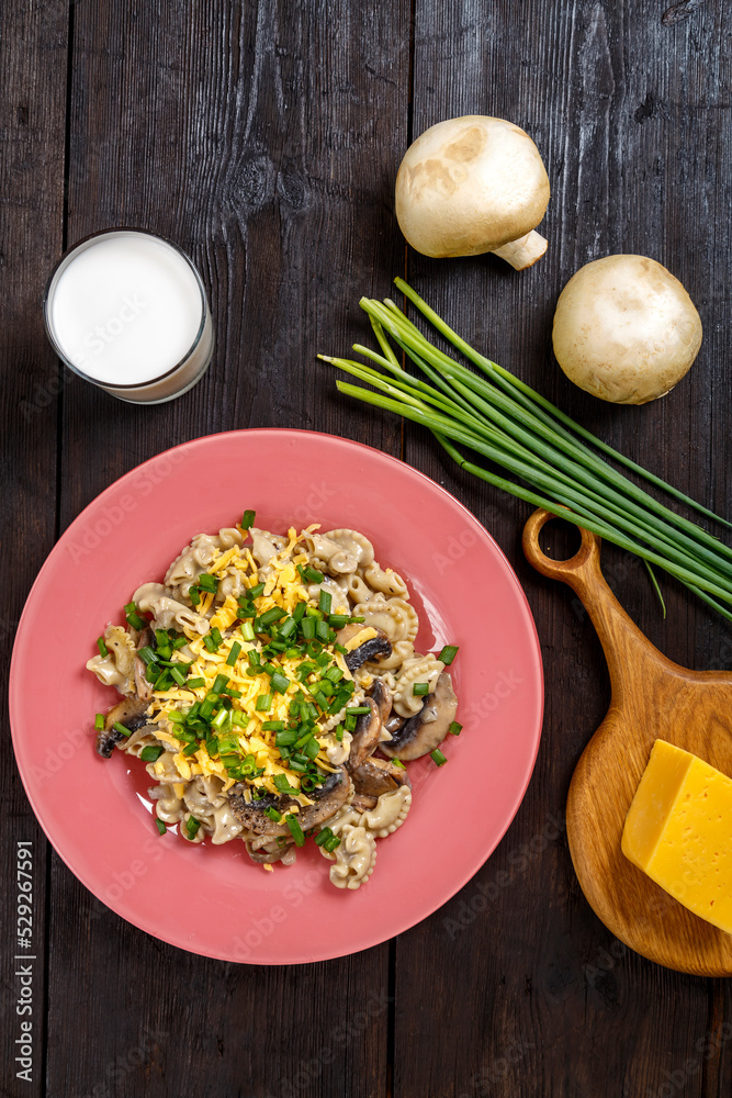 Pasta in a creamy sauce with mushrooms in a plate on the table next to a glass of cream, cheese on a wooden board, green onions and champignons top view.
