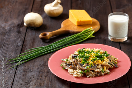 Pasta in a creamy sauce with mushrooms in a plate on a dark table next to a glass of cream, cheese on a wooden board, green onions and mushrooms.