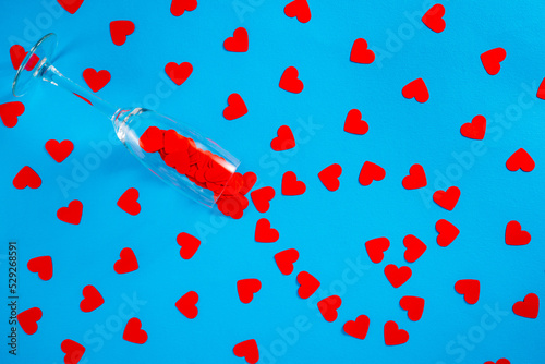 Wineglass and pattern of paper red hearts on blue screen background. Top view of heart shaped confetti arranged in a circle around a glass. Screensaver mockup for Valentine's day, holiday, party.