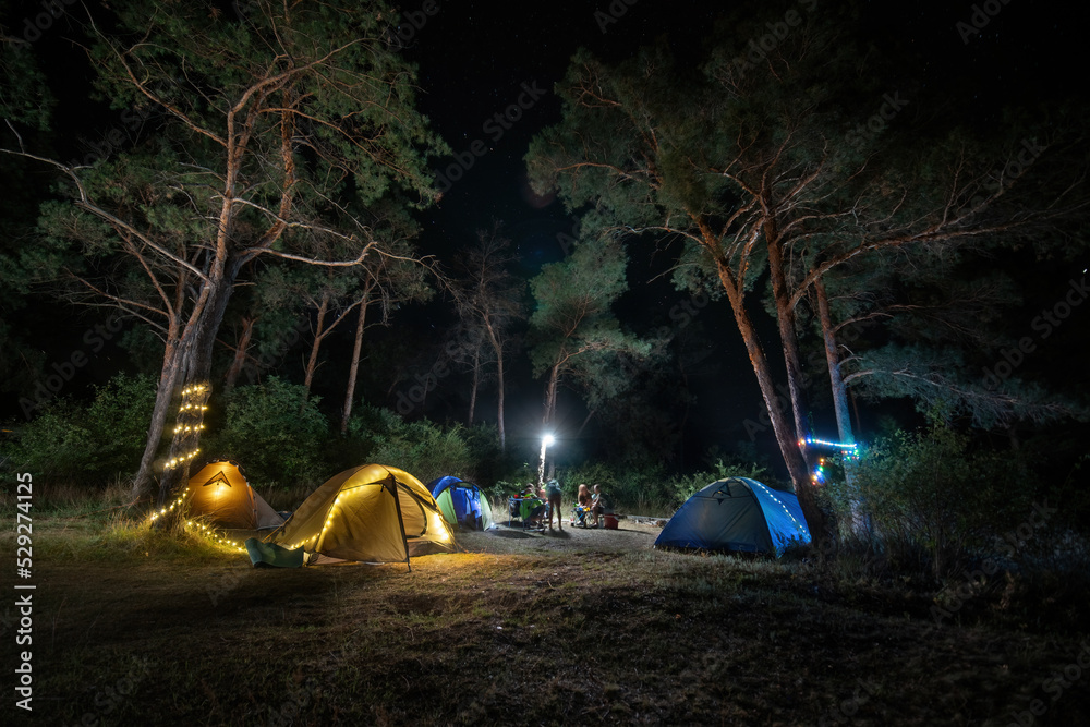 Camping in forest at night. garlands, light bulbs shine between pine trees and tents, people sitting opposite each other near illuminated tents under beautiful evening starry sky