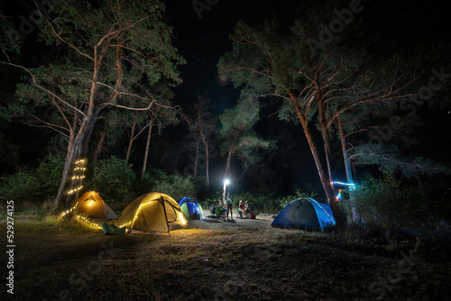 Obraz na płótnie Camping in forest at night. garlands, light bulbs shine between pine trees and tents, people sitting opposite each other near illuminated tents under beautiful evening starry sky w salonie