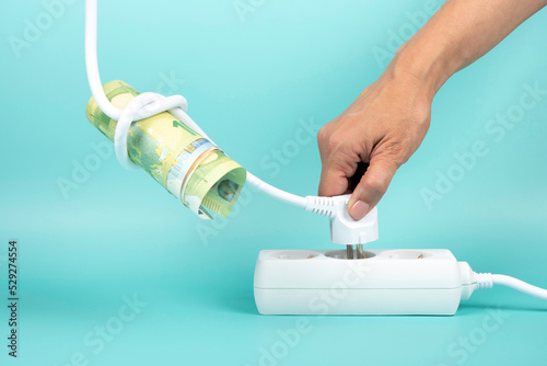 Woman hand holding electric power plug with Euro banknote on cord and plugging it in power strip extension cord on blue background. Electricity cost, electric prices increase, expensive energy concept photo