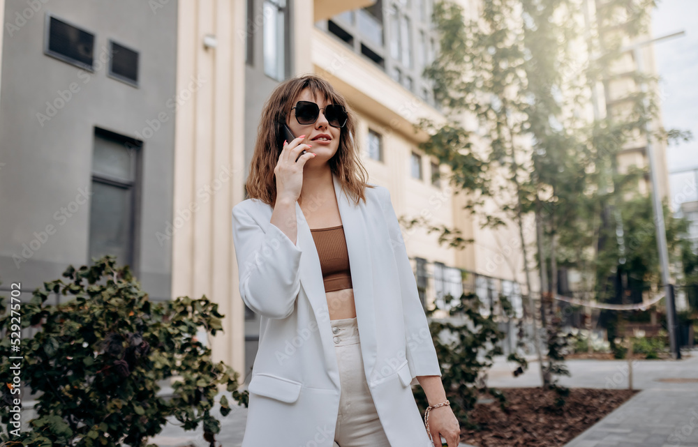 Smiling businesswoman in white suit talking phone during walking in city with modern architecture