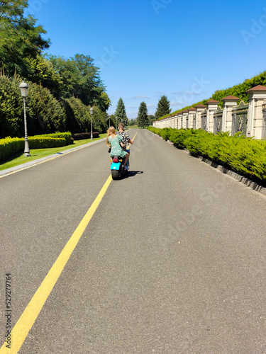 A boy and a girl are riding an electric scooter in perspective on a long road in a city park during the day