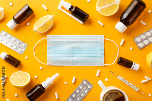 Seasonal diseases concept. Top view photo of face mask medicines spray and syrup bottles pills blisters thermometer cut lemon and cup of tea on isolated orange background