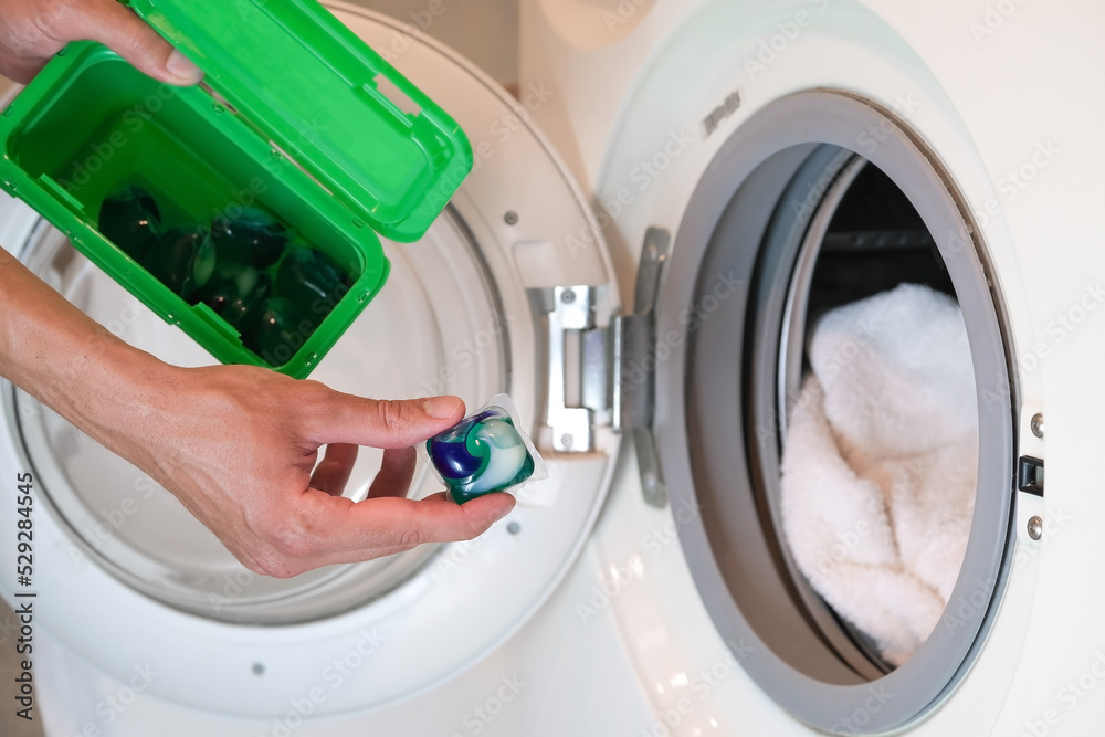 The hand holds a laundry detergent capsule and starts putting it into the open washing machine The hand holds a laundry detergent capsule and starts putting it into the open washing machine